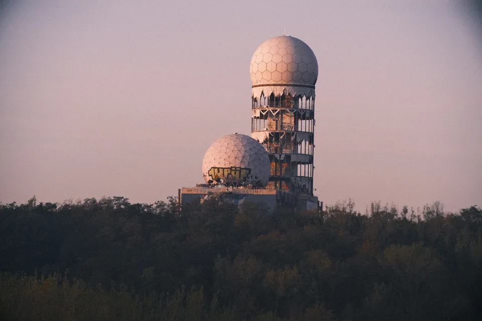 Zwei kuppelförmige Türme stehen über einer dunklen Waldlinie unter einem blassen Abendhimmel.