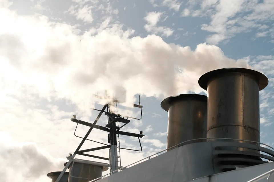 Zwei große Schornsteine auf einem Schiff stoßen weißen Rauch in den bewölkten Himmel.