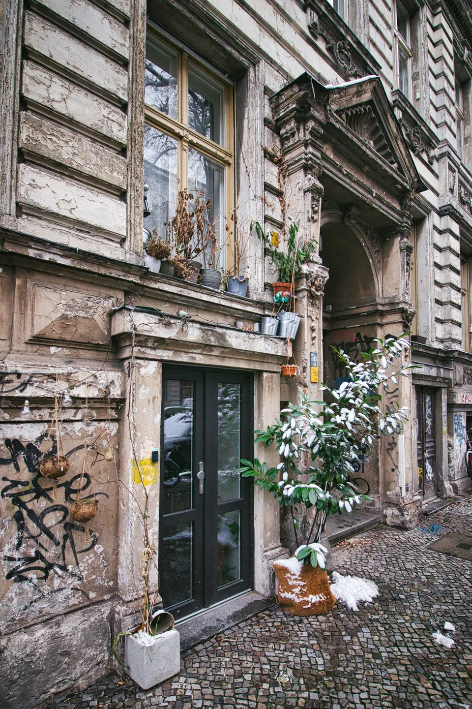 Eine alte, verwitterte Hausfassade mit Pflanzen in Töpfen, Graffiti und einer Tür im Schnee.