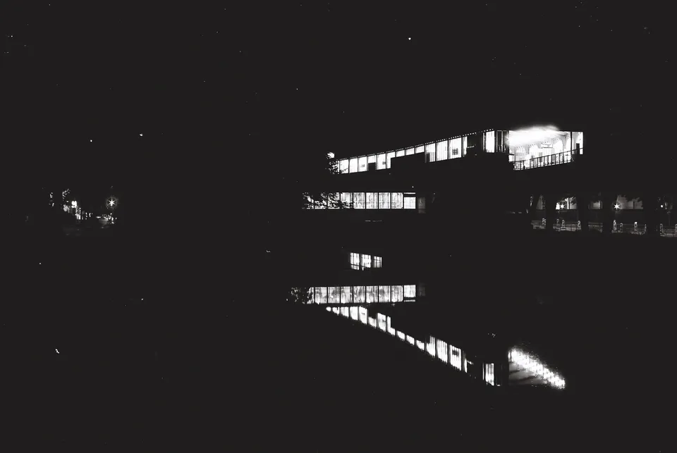 Die hell erleuchteten Fenster eines Gebäudes spiegeln sich in dunklem Wasser bei Nacht.