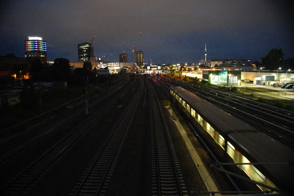 Blick auf Gleise und einen beleuchteten Zug in einer Stadt bei Nacht.