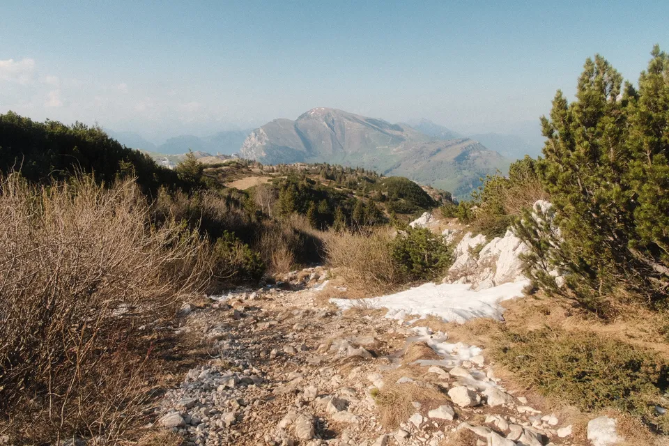 Ein felsiger Wanderpfad führt durch trockenes Gebüsch und Nadelbäume zu einer bergigen Landschaft.