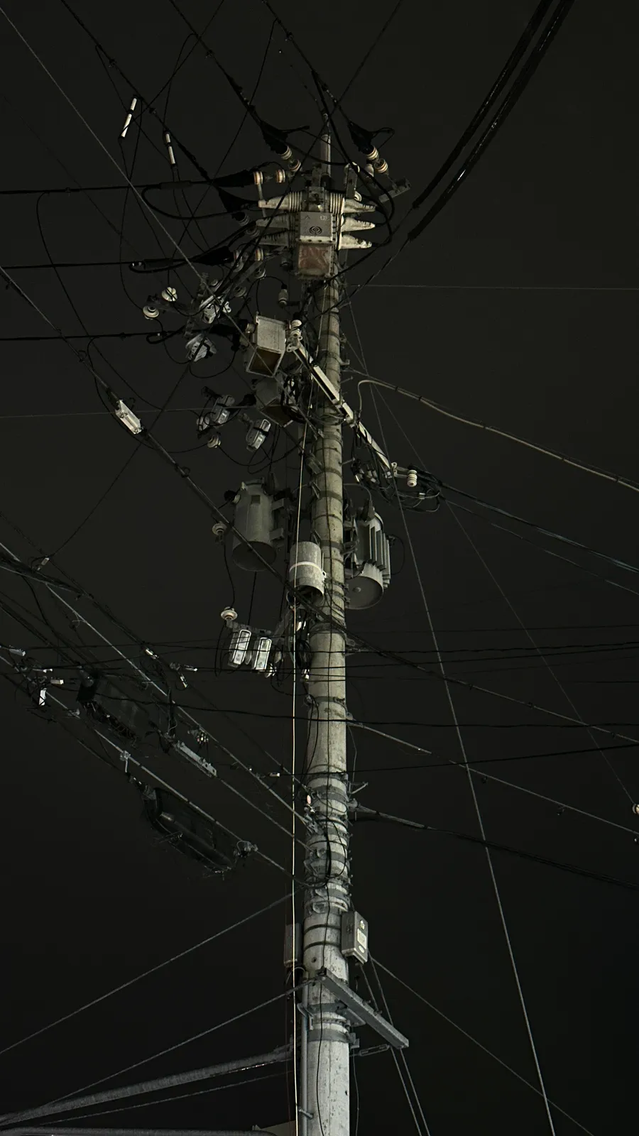 Ein komplexes Geflecht aus Kabeln und Transformatoren an einem Strommast vor dunklem Hintergrund.