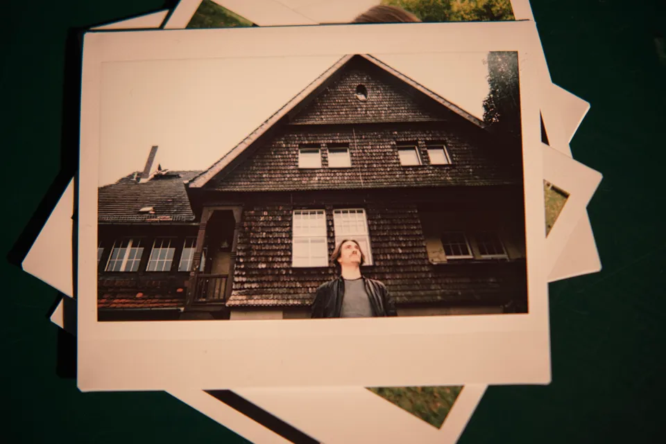 Eine Person blickt nach oben vor einem hölzernen Haus auf einem Polaroid-Foto.