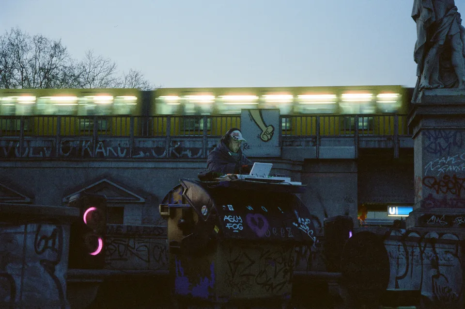 Eine Person sitzt vor einem Laptop auf einer mit Graffiti verzierten Mauer vor einer beleuchteten Brücke.