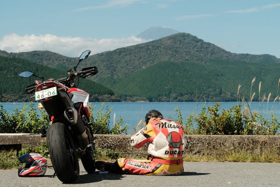 Eine Person in Motorradbekleidung sitzt neben einem Motorrad an einem See mit Bergen im Hintergrund.
