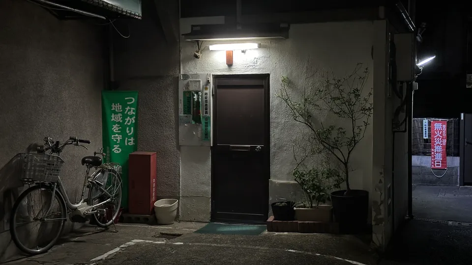 Eine beleuchtete Tür in einer schmalen Gasse bei Nacht mit einem abgestellten Fahrrad.