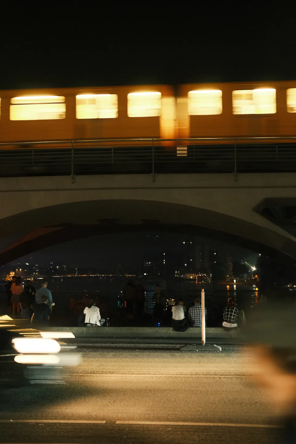 Eine beleuchtete Brücke über einer Straße mit Menschen am Wasser in der Nacht.