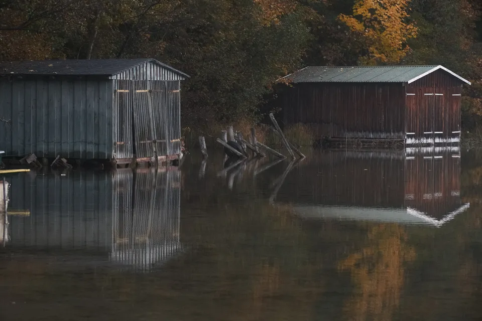 Zwei hölzerne Bootshäuser spiegeln sich im ruhigen Wasser vor einer herbstlichen Waldkulisse.
