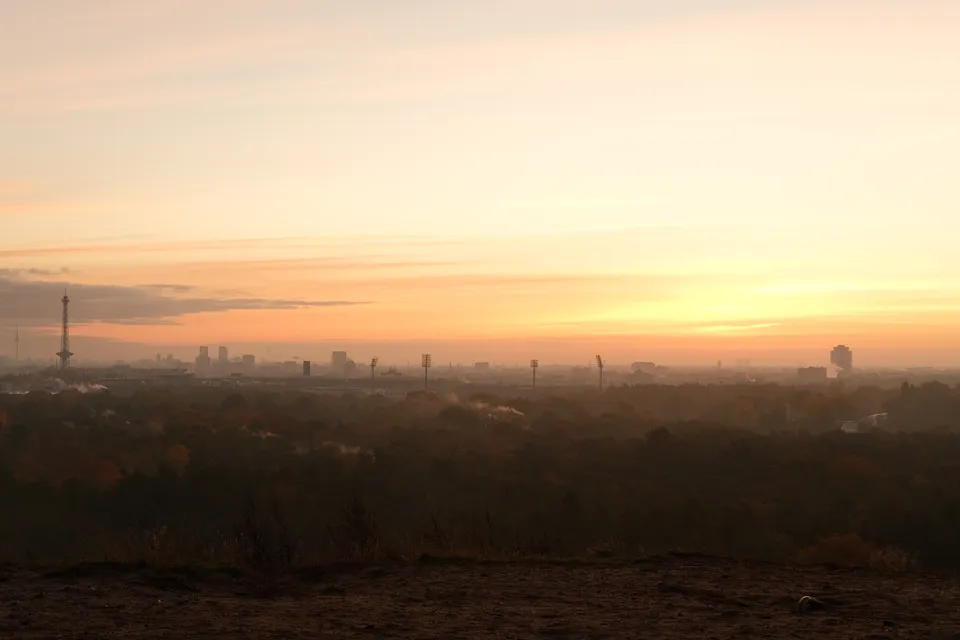 Eine weite Stadtansicht bei Sonnenuntergang mit Silhouetten von Gebäuden und Türmen vor einem orangefarbenen Himmel.