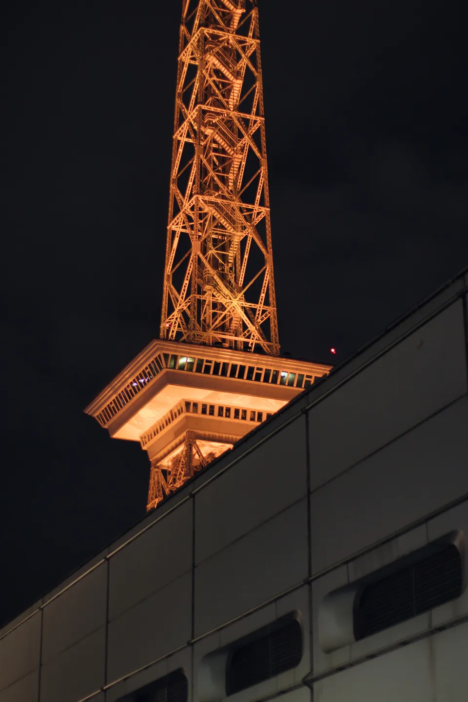 Ein beleuchteter Gittermastturm ragt über das Dach eines modernen Gebäudes in die Nacht.