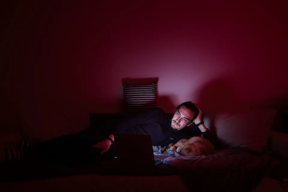 Eine Person liegt in einem dunklen Raum mit einem Hund und einem Laptop auf dem Bett.