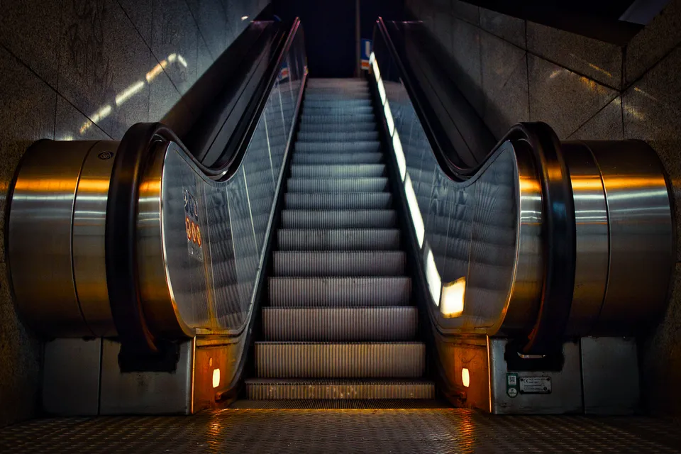 Eine beleuchtete Rolltreppe führt in der Nacht nach oben zwischen metallischen Wänden.