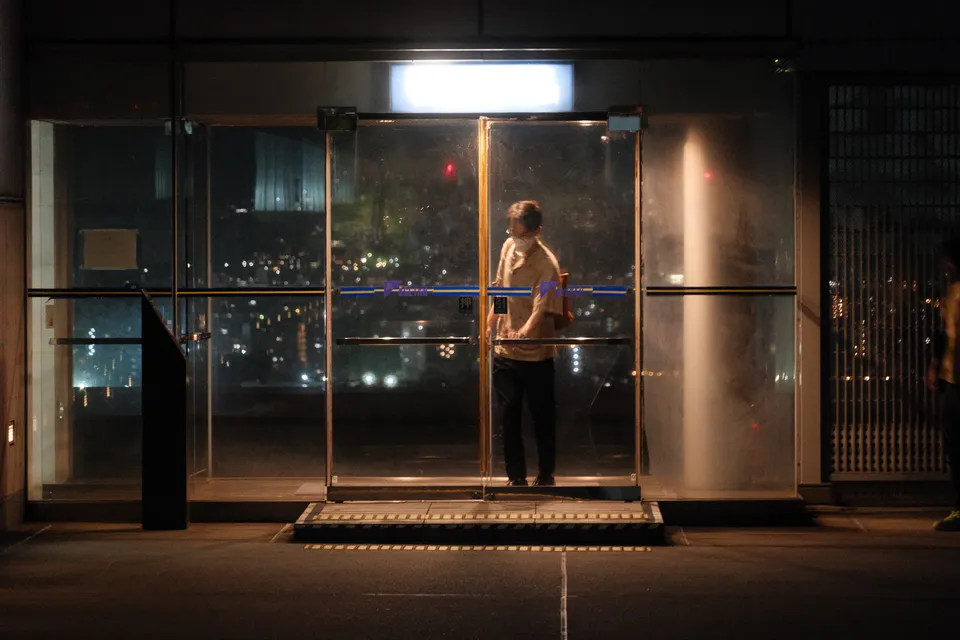 Eine Person steht in einem beleuchteten Gebäude hinter einer großen Glasfront mit Blick auf eine nächtliche Stadt.