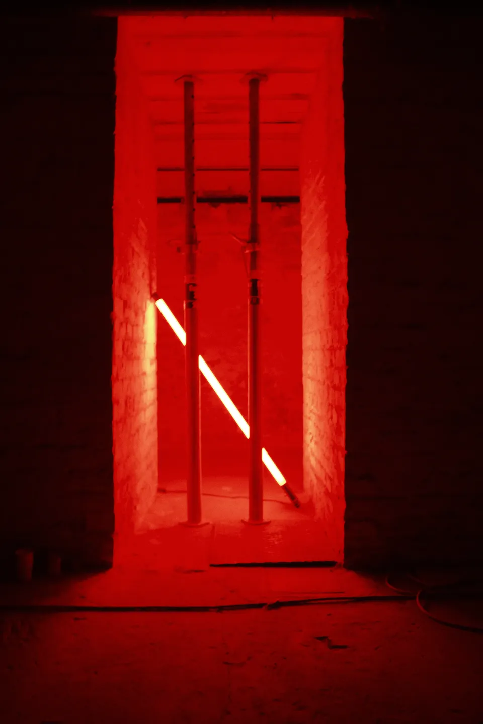 Ein dunkler Raum mit einer durch eine Tür beleuchteten, roten Nische, in der eine diagonale Lichtröhre zu sehen ist.
