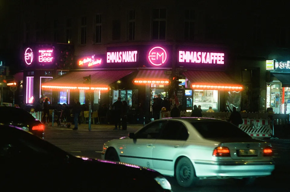 Eine nächtliche Straßenszene mit einem hell erleuchteten Laden und Personen auf dem Gehweg.