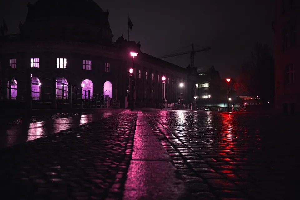 Eine regennasse Kopfsteinpflasterstraße spiegelt die pinken und roten Lichter einer beleuchteten Architektur wider.