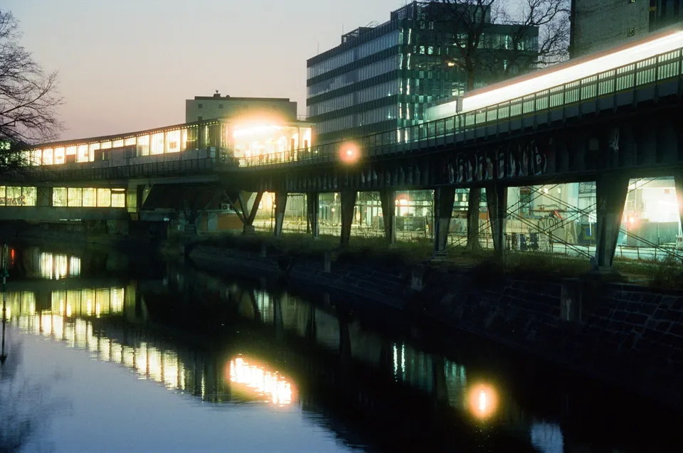 Eine hell beleuchtete Bahnbrücke führt über einen Fluss und spiegelt sich im Wasser wider.
