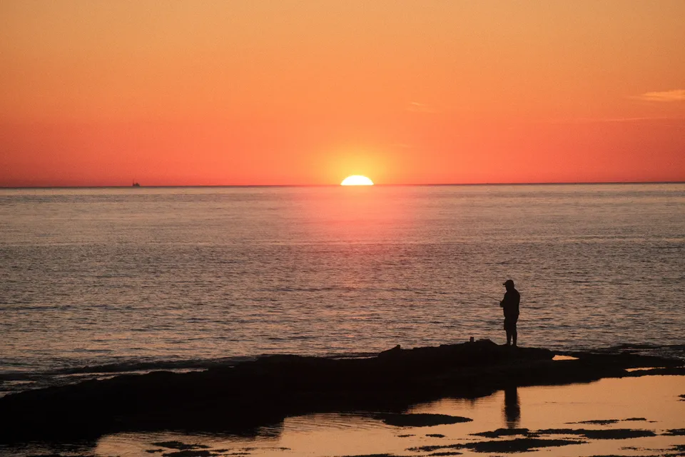 Eine Person steht auf einem Felsen am Ufer und betrachtet den Sonnenuntergang über dem Meer.