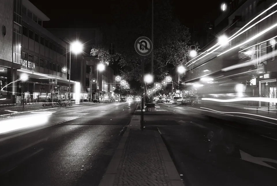 Langzeitbelichtung einer nächtlichen Straße mit vorbeifahrenden Fahrzeugen und beleuchteten Gebäuden.