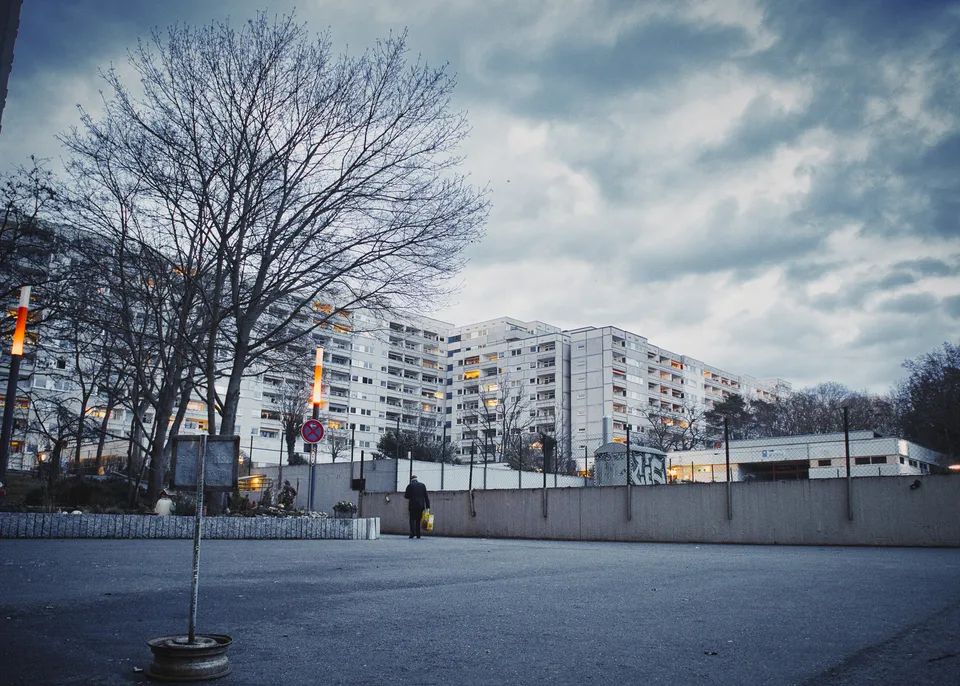 Große Wohnblöcke hinter einem kahlen Baum und einer niedrigen Mauer unter einem bewölkten Himmel.