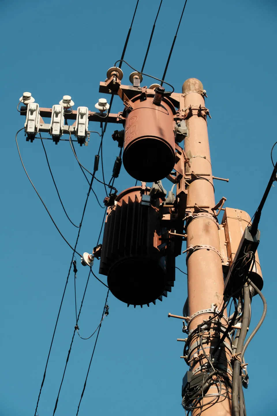 Ein rostfarbener Strommast mit Transformatoren und Kabeln vor einem klaren blauen Himmel.