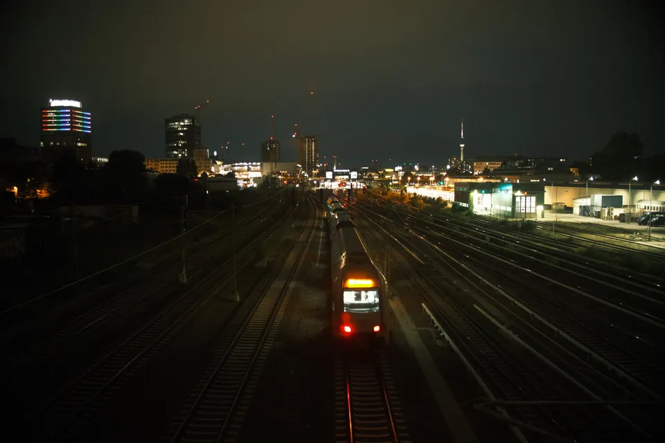 Ein Zug fährt nachts durch ein weitläufiges Bahngelände mit beleuchteten Stadtgebäuden im Hintergrund.