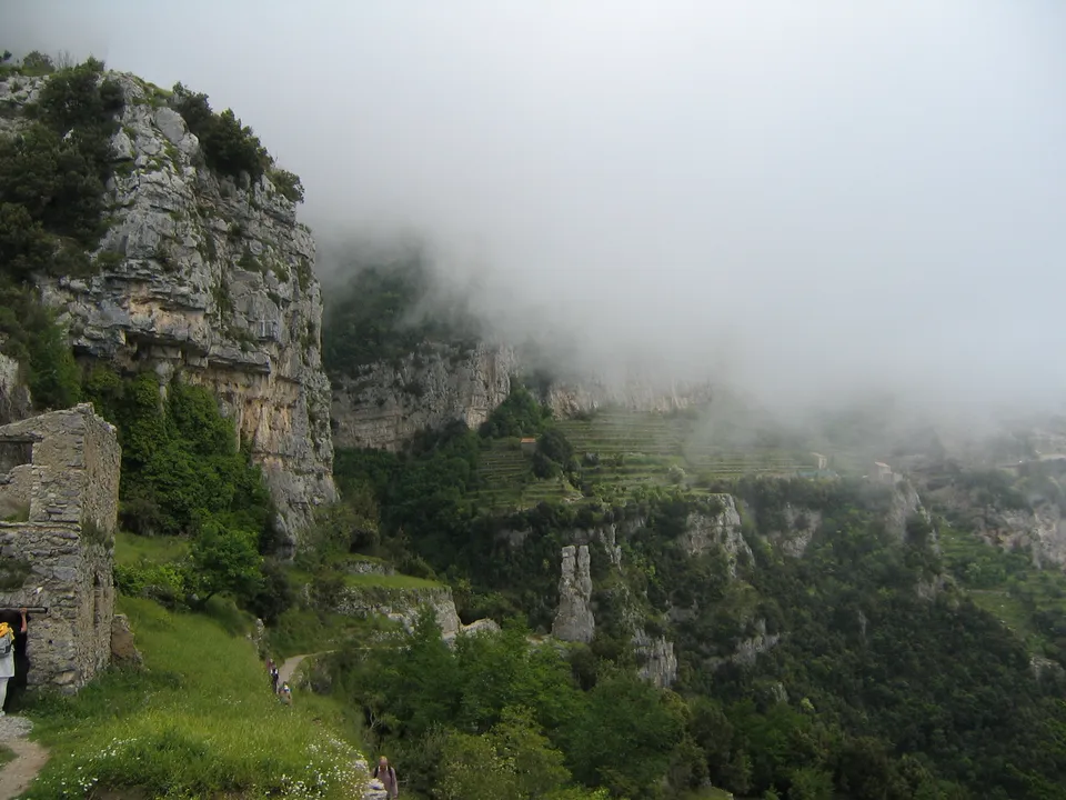 Eine neblige Gebirgslandschaft mit Felswänden, grünen Terrassenfeldern und Wanderern auf einem Pfad.