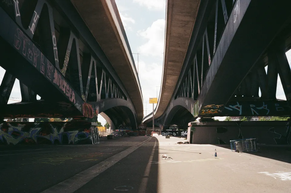 Zwei große Metallbrücken ragen über eine sonnenbeschienene Straße mit Graffiti an den Pfeilern.