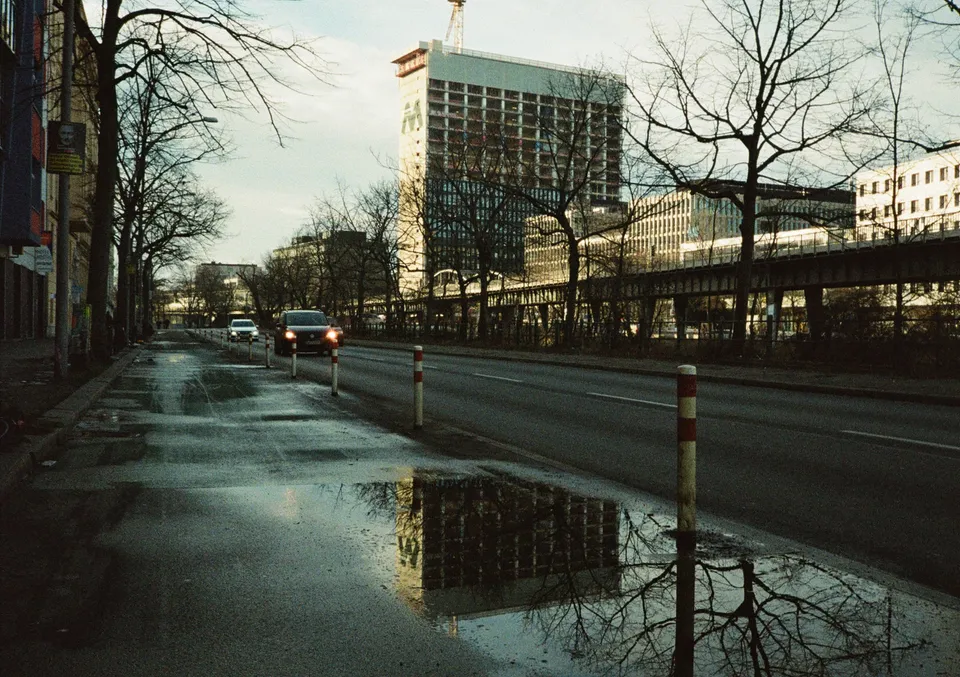 Eine nasse Straße mit Pfützen reflektiert ein großes Gebäude im Hintergrund.