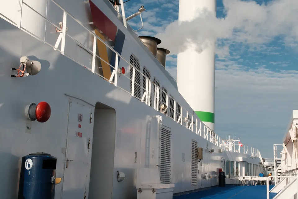 Nahaufnahme der weißen Außenwand eines Schiffes mit Geländern, Lüftungsgittern und einem Schornstein im Hintergrund.
