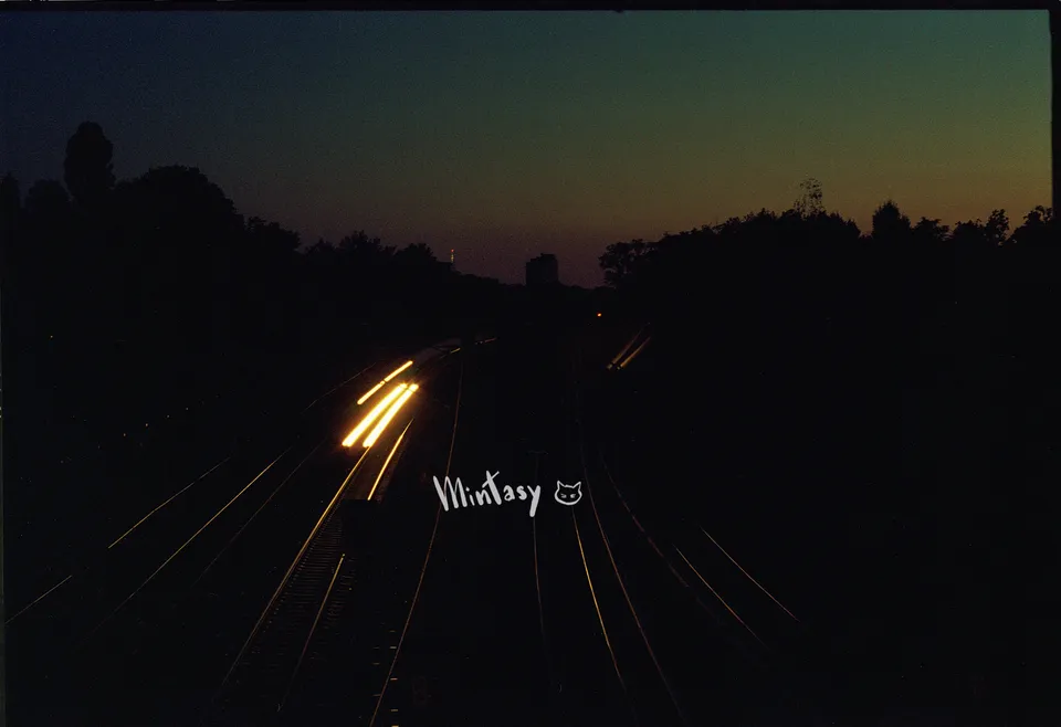 Lange Belichtung von Lichtspuren auf Bahngleisen bei Dämmerung vor einer dunklen Silhouette.