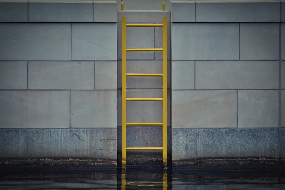 Eine gelbe Metallleiter ist an einer grau strukturierten Betonmauer über dem Wasser befestigt.