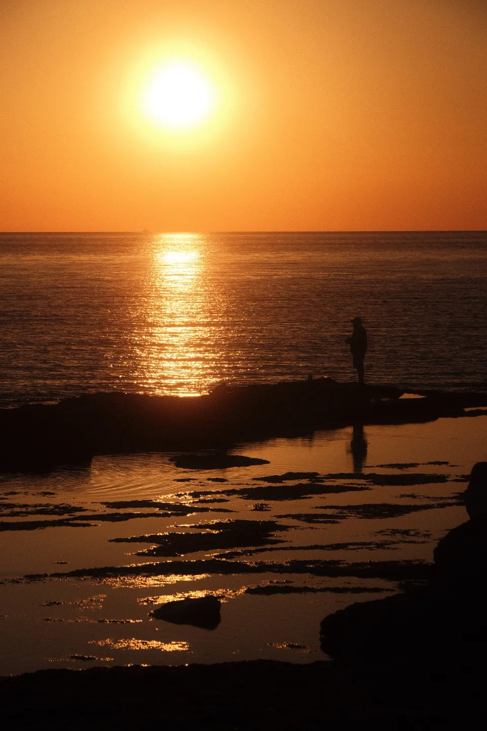 Eine Person steht am Ufer und blickt auf den glitzernden Sonnenuntergang über dem Meer.