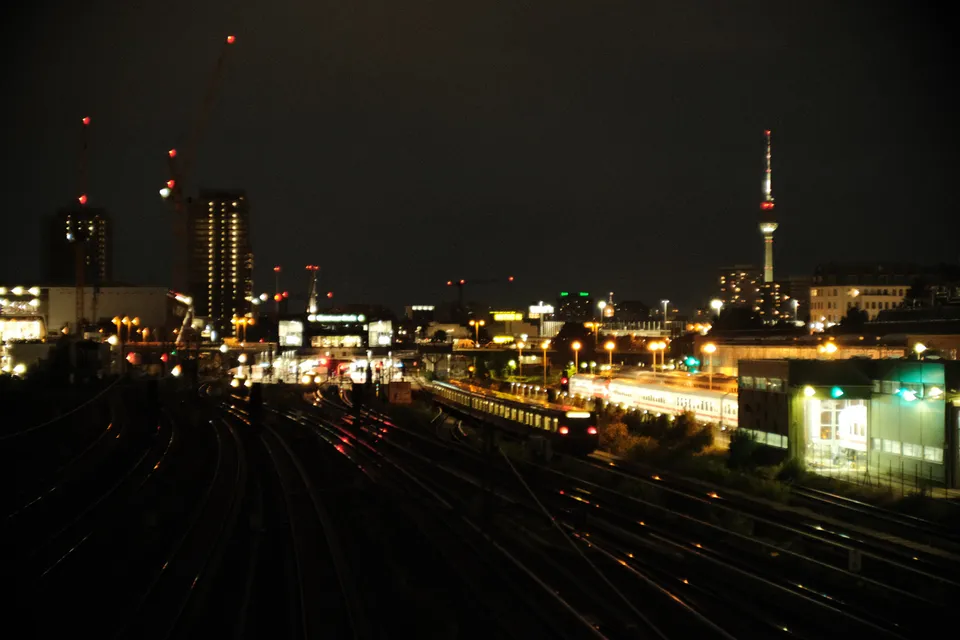 Eine nächtliche Stadtansicht mit beleuchteten Bahngleisen, Gebäuden und einem hohen Turm im Hintergrund.