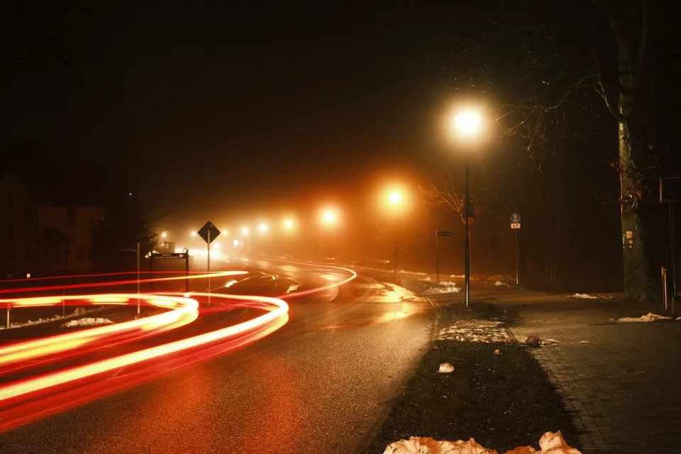 Langzeitbelichtung zeigt rote Lichtspuren eines Fahrzeugs auf einer dunklen, verschneiten Straße bei Nacht.