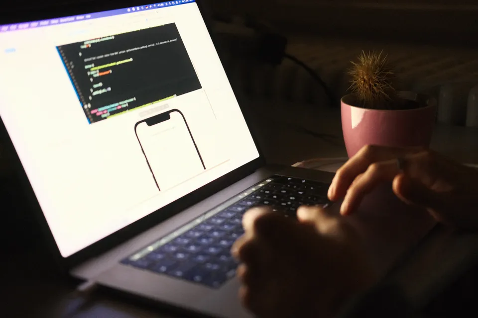 Hände einer Person tippen auf die Tastatur eines Laptops, der Programmiercode anzeigt.