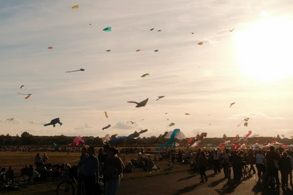 Viele bunte Drachen schweben im Himmel über einer Menschenmenge auf einem Feld während des Sonnenuntergangs.