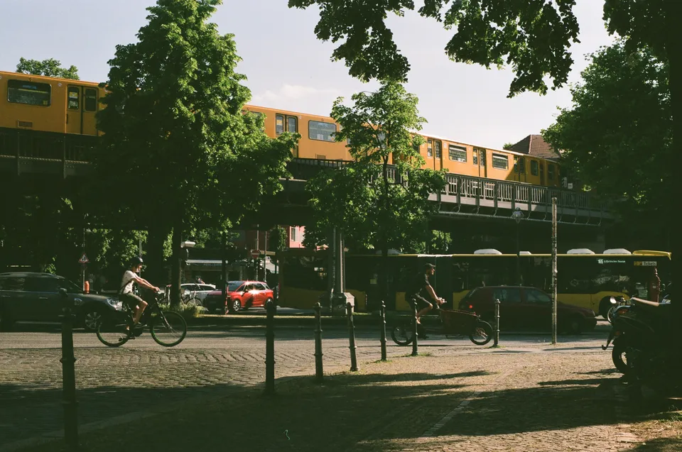Zwei Personen fahren mit Fahrrädern an einer Straße mit einer gelben Hochbahn vorbei.