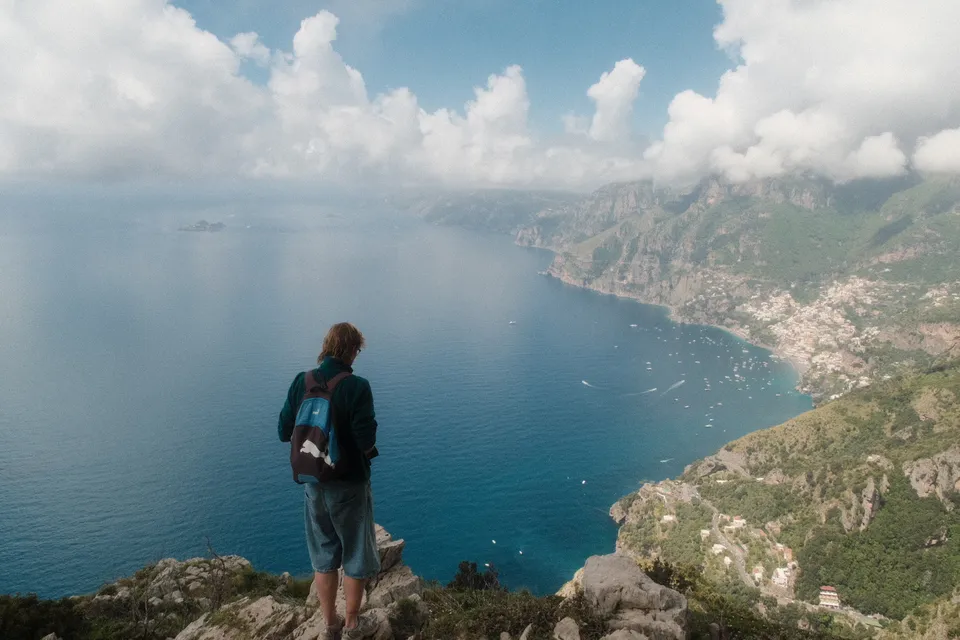 Eine Person steht auf einem Felsen und blickt auf eine Küstenlandschaft mit Bergen und Meer.