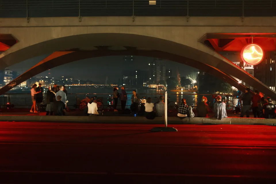 Eine Gruppe von Menschen sitzt und steht unter einer beleuchteten Brücke am Wasser bei Nacht.