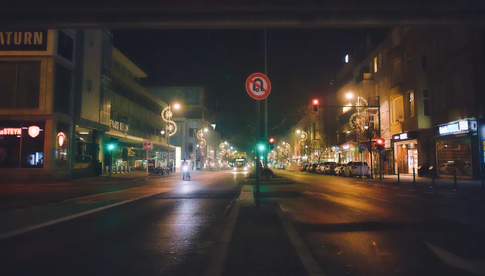 Eine beleuchtete Straße bei Nacht mit Gebäuden, Ampeln und einer Person, die auf dem Gehweg steht.