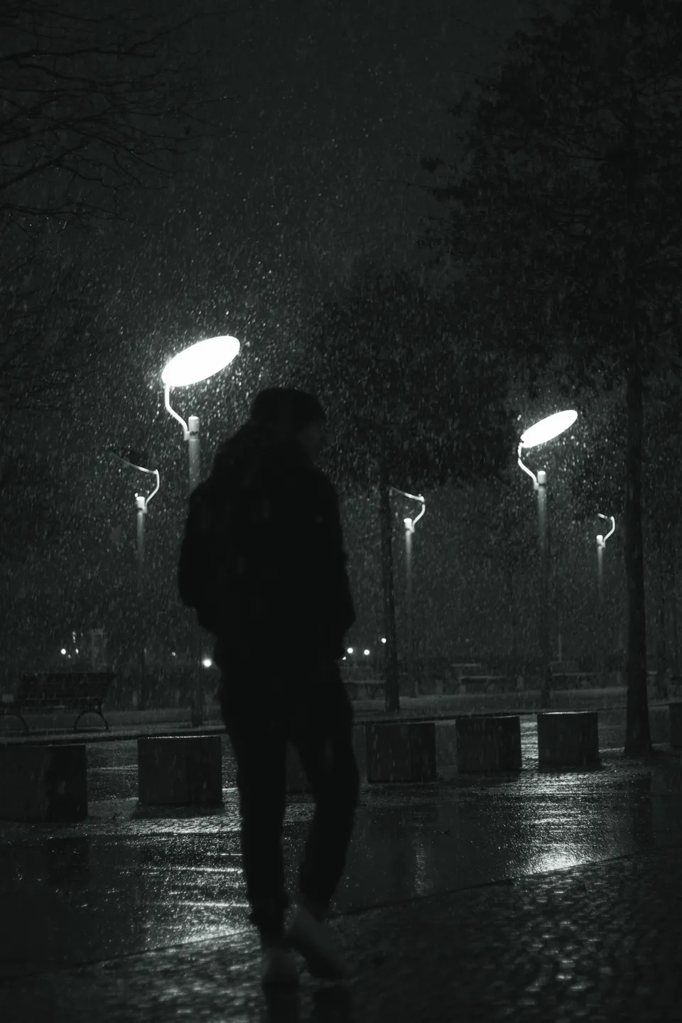 Eine dunkle Silhouette einer Person geht bei nächtlichem Regen unter Straßenlaternen.